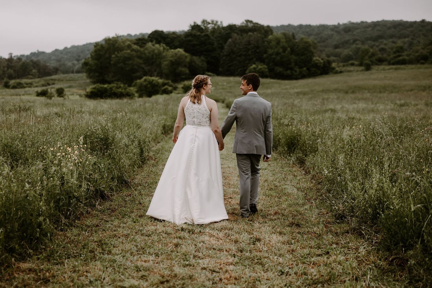 Bride and groom walking hand in hand through green meadow path during outdoor wedding portrait session.