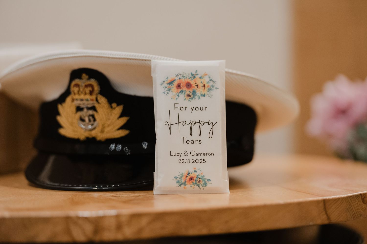 A military cap with an emblem sits next to a delicate floral card reading 'for your happy tears' on a wooden surface.