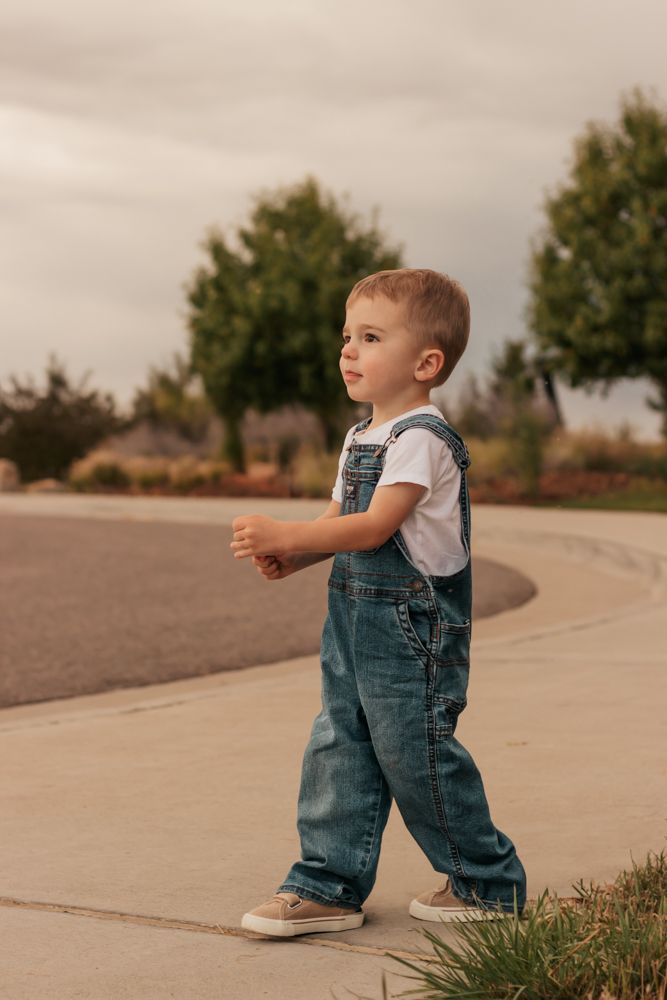 A toddler in denim overalls and white shirt walks along a curved sidewalk on a cloudy day.| Westminster Colorado Family Photographer