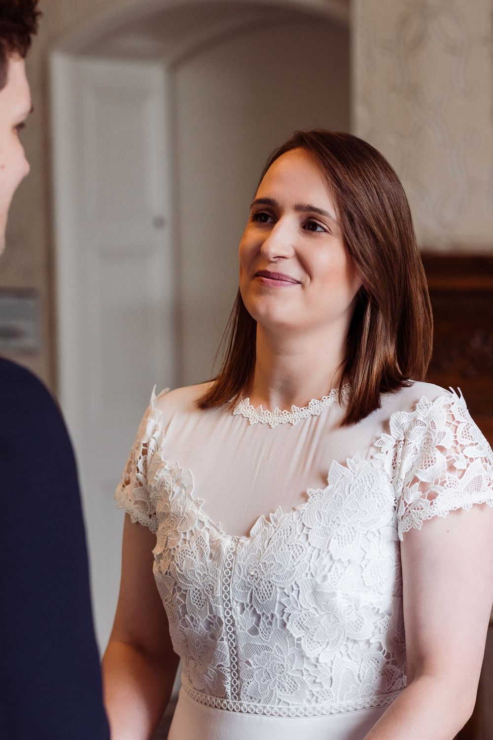 A bride in a delicate white lace wedding dress smiles warmly during an intimate indoor ceremony.