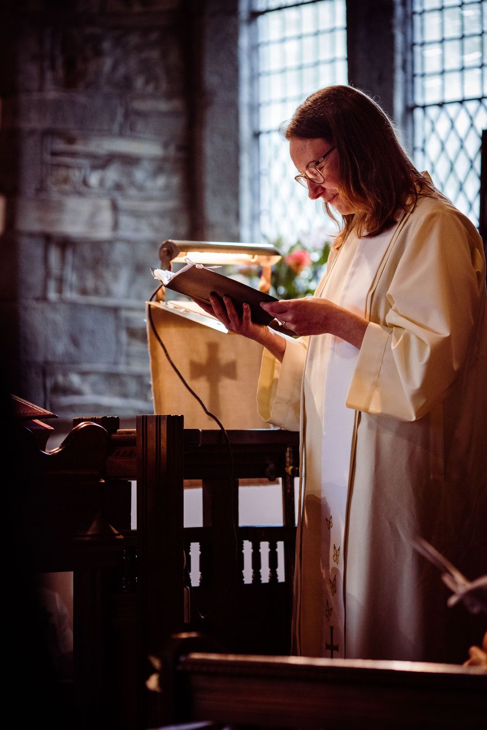 A person in white robes reads from a religious text at a wooden church pulpit in atmospheric lighting.