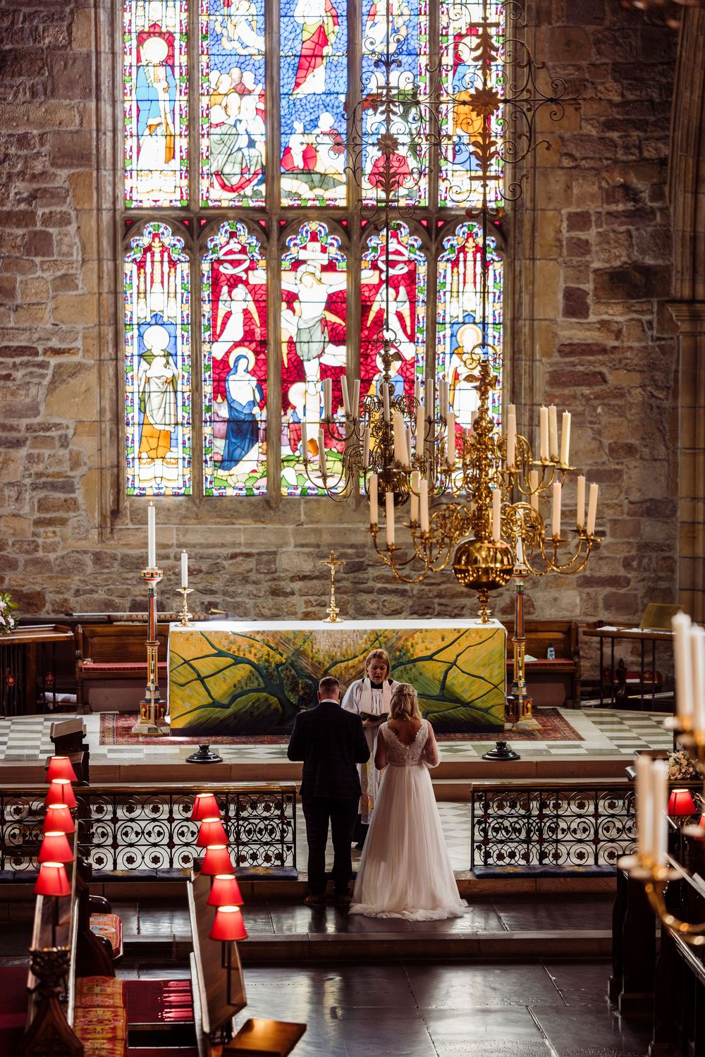 Beautiful stained glass windows illuminate a church altar during a wedding ceremony with red and white decor.