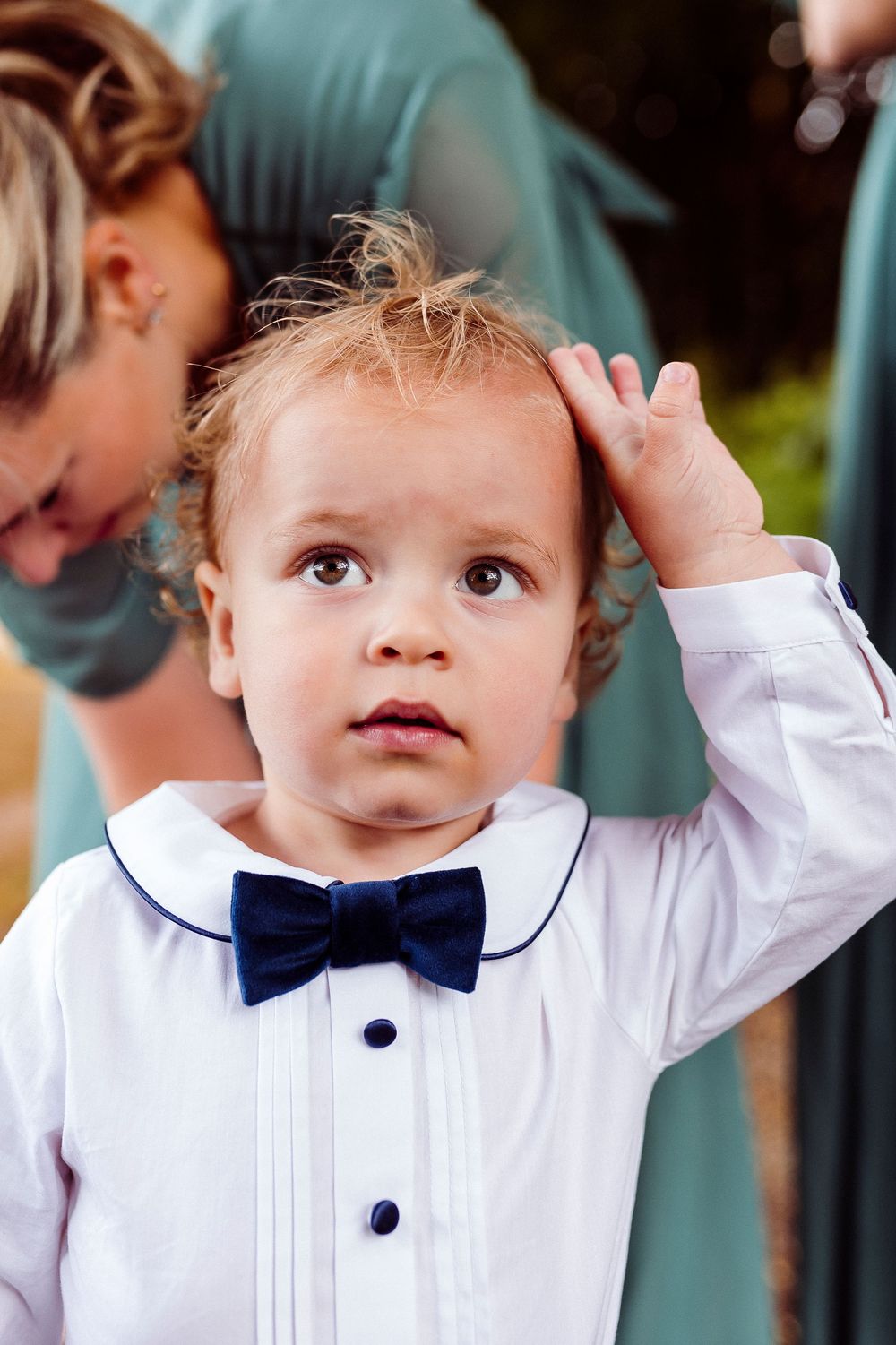 A toddler in a white dress shirt and navy bow tie looks up with an adorable expression while being held.