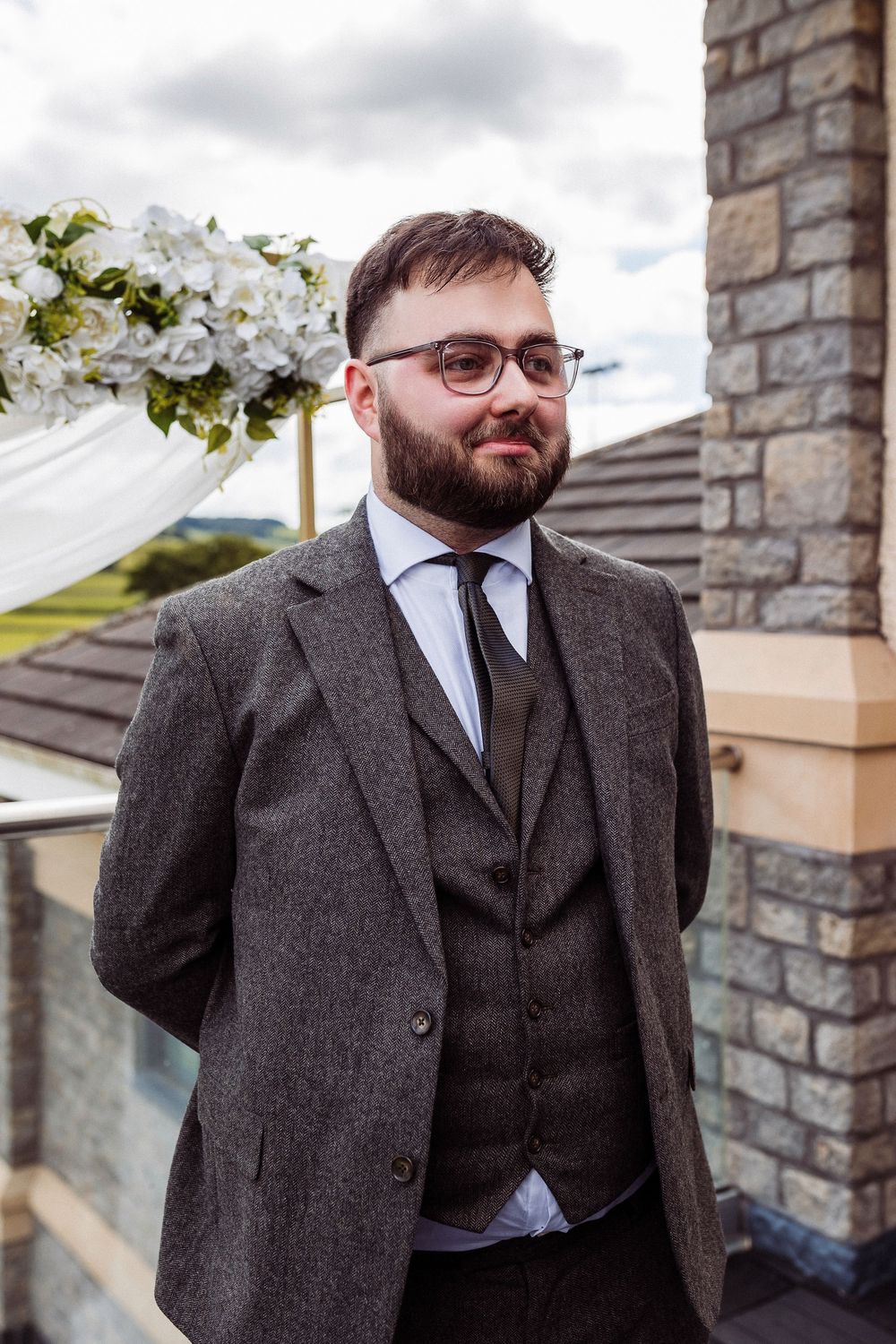 A person in a dark gray wool suit and glasses stands against a stone wall backdrop outdoors.