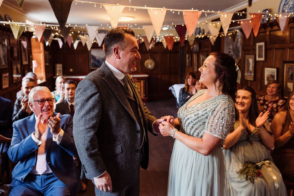 A couple shares a joyful moment during their wedding ceremony while guests applaud in a rustic venue with bunting decorations.
