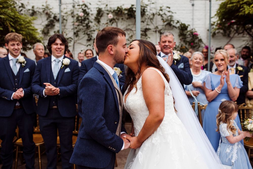 A romantic kiss between newlyweds surrounded by guests at an outdoor wedding celebration.