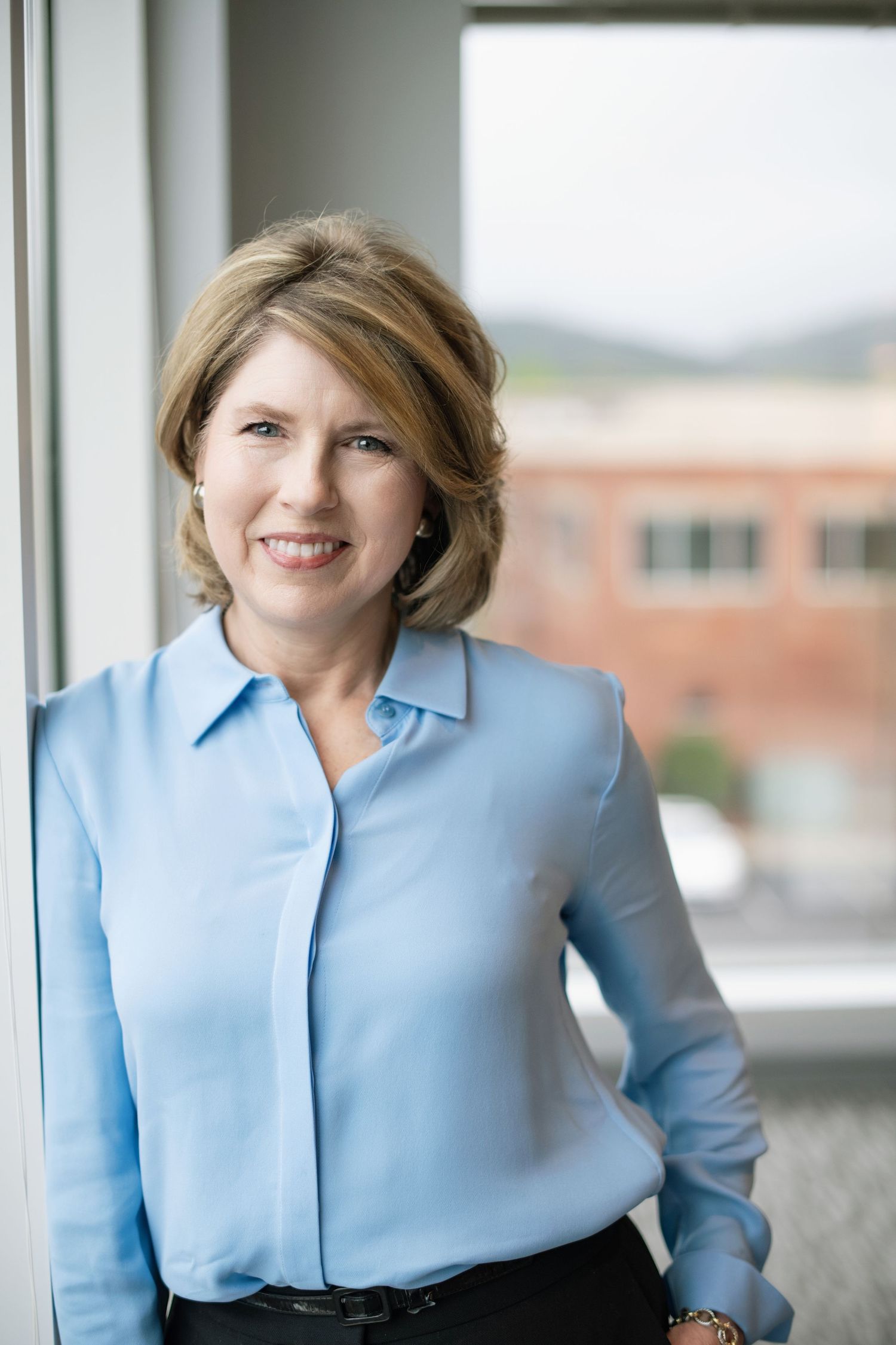 Professional wearing a light blue collared shirt poses against a bright window background in an office setting.