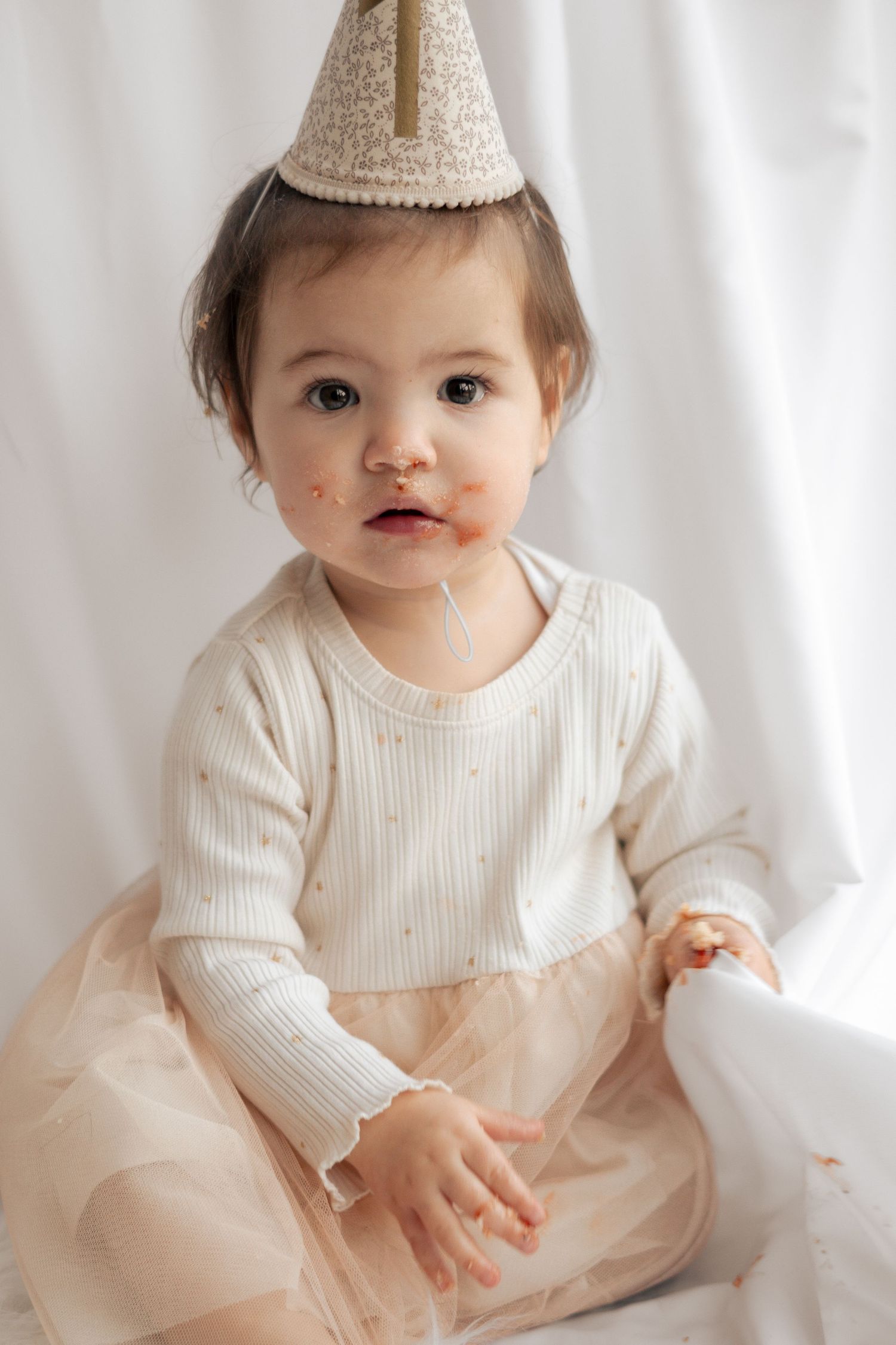 Baby wearing a white dress and sparkly party hat sits against white backdrop during first birthday photoshoot.Photography in Buckinghamshire