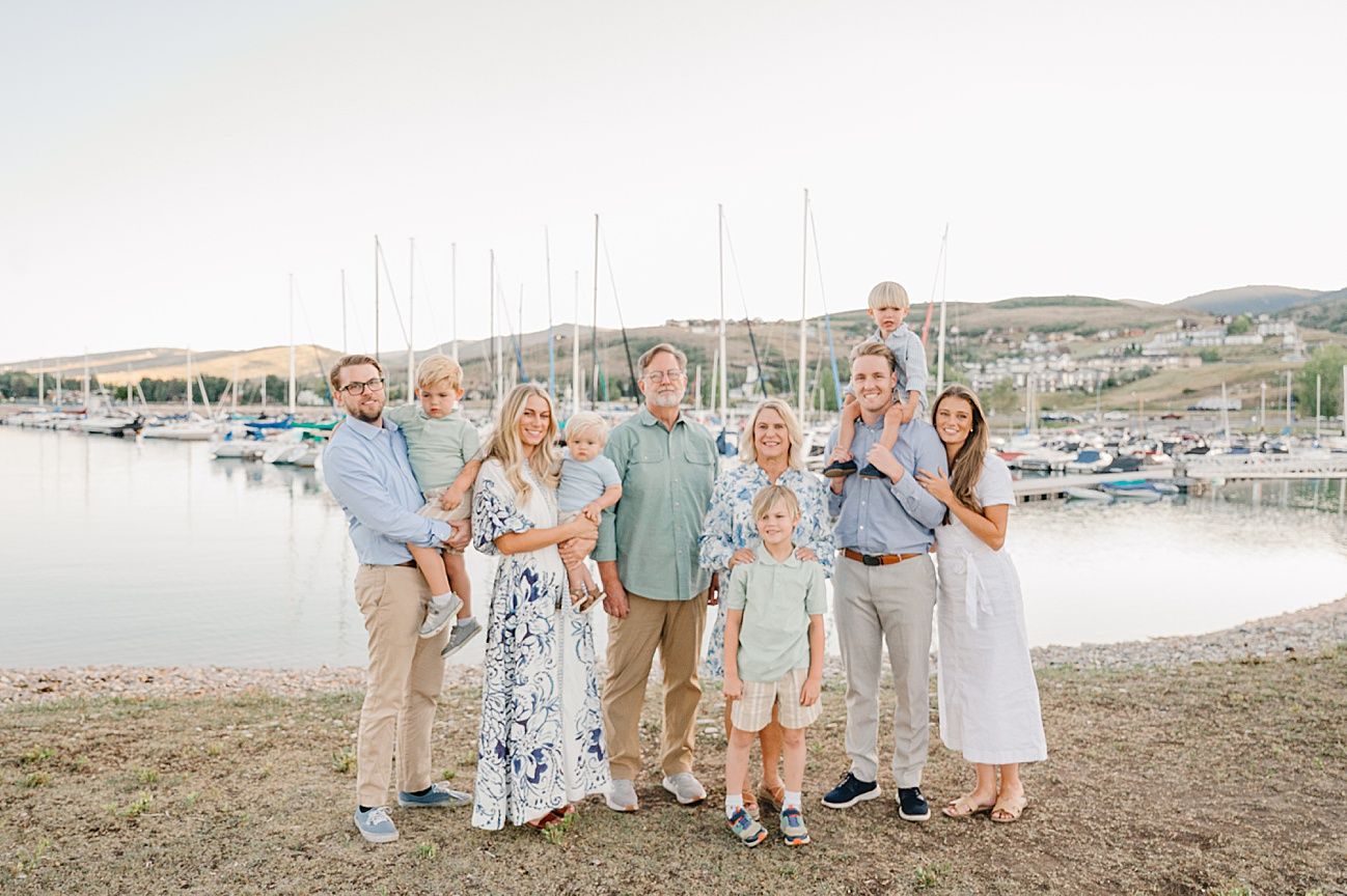 Family group wearing coordinated blue and neutral outfits poses together on a beach with boats and marina in background.