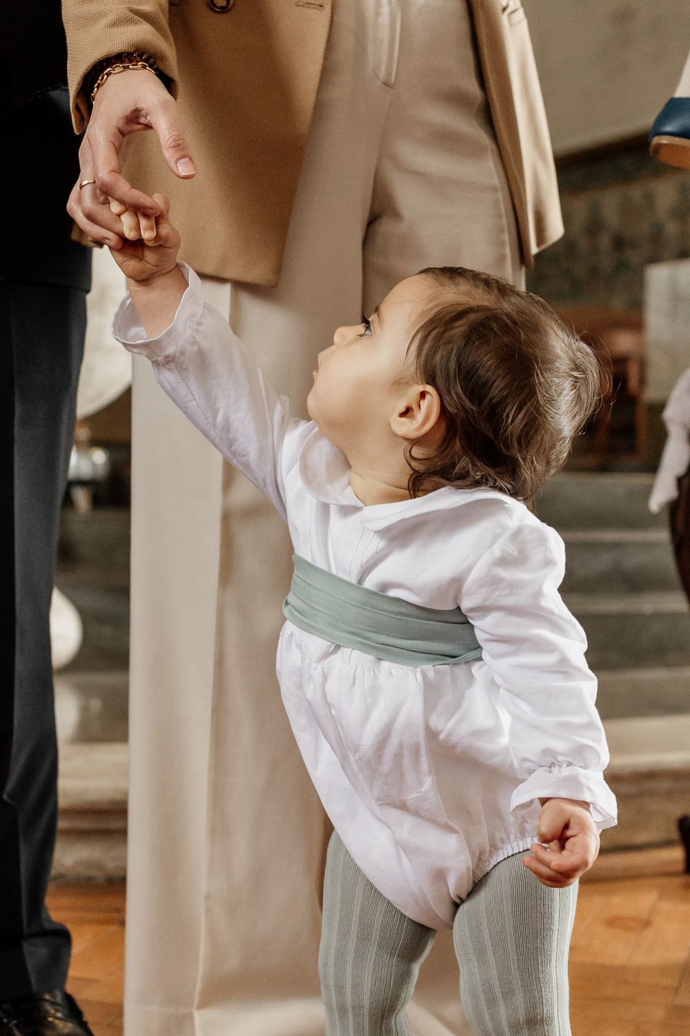 A small child in white shirt and gray tights reaches up while holding hands with a caregiver wearing beige clothing.