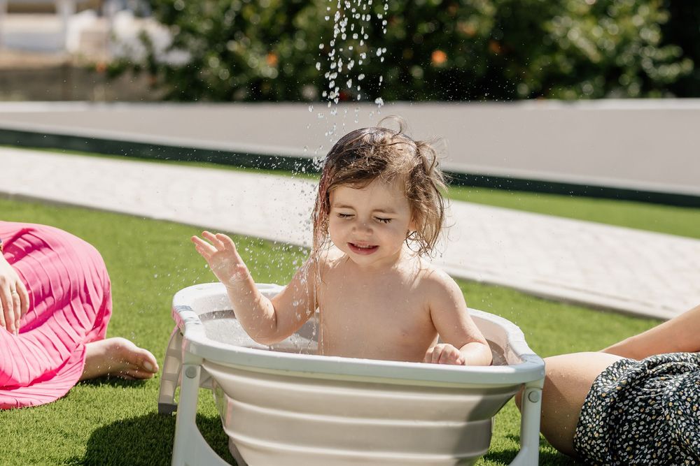 A child splashes and plays in a white washtub outdoors on a sunny day.