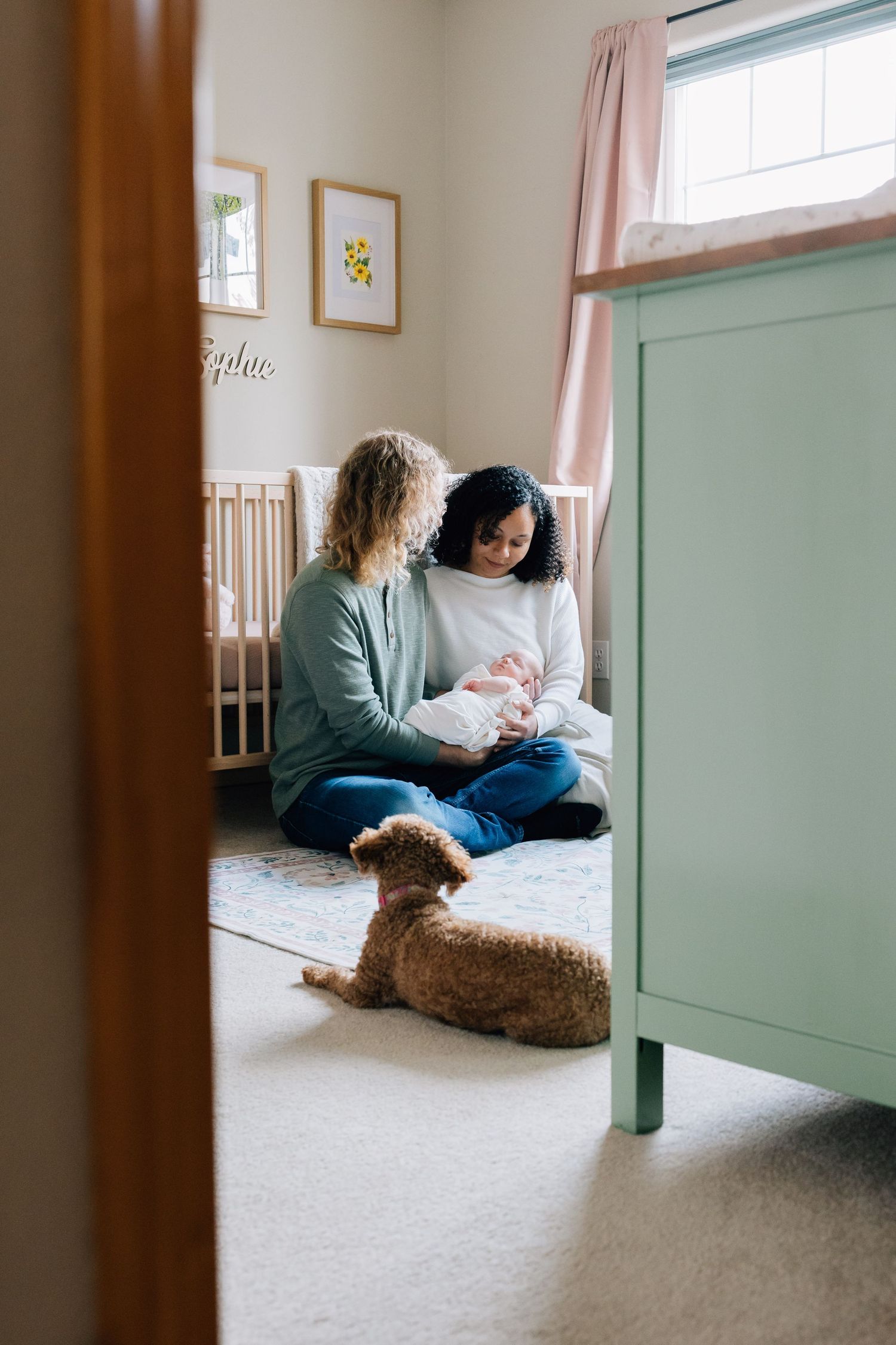 Two mothers sitting on nursery floor with newborn baby and fluffy brown dog.