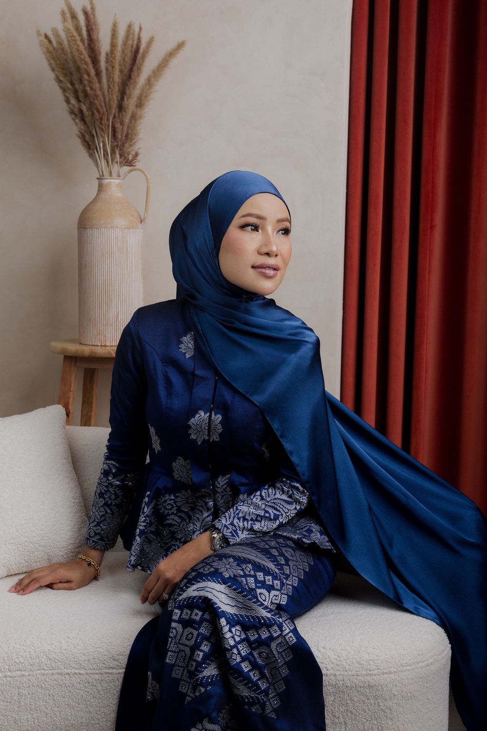 A person wearing a navy blue hijab and embroidered dress poses elegantly against a white wall with red curtains.