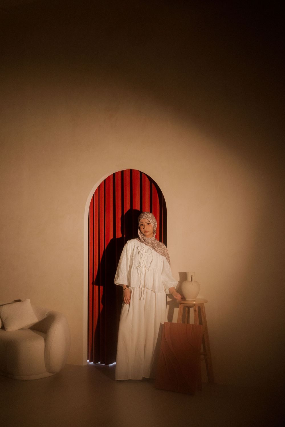 Woman in white dress standing in front of red arched doorway with warm beige walls and wooden stool.