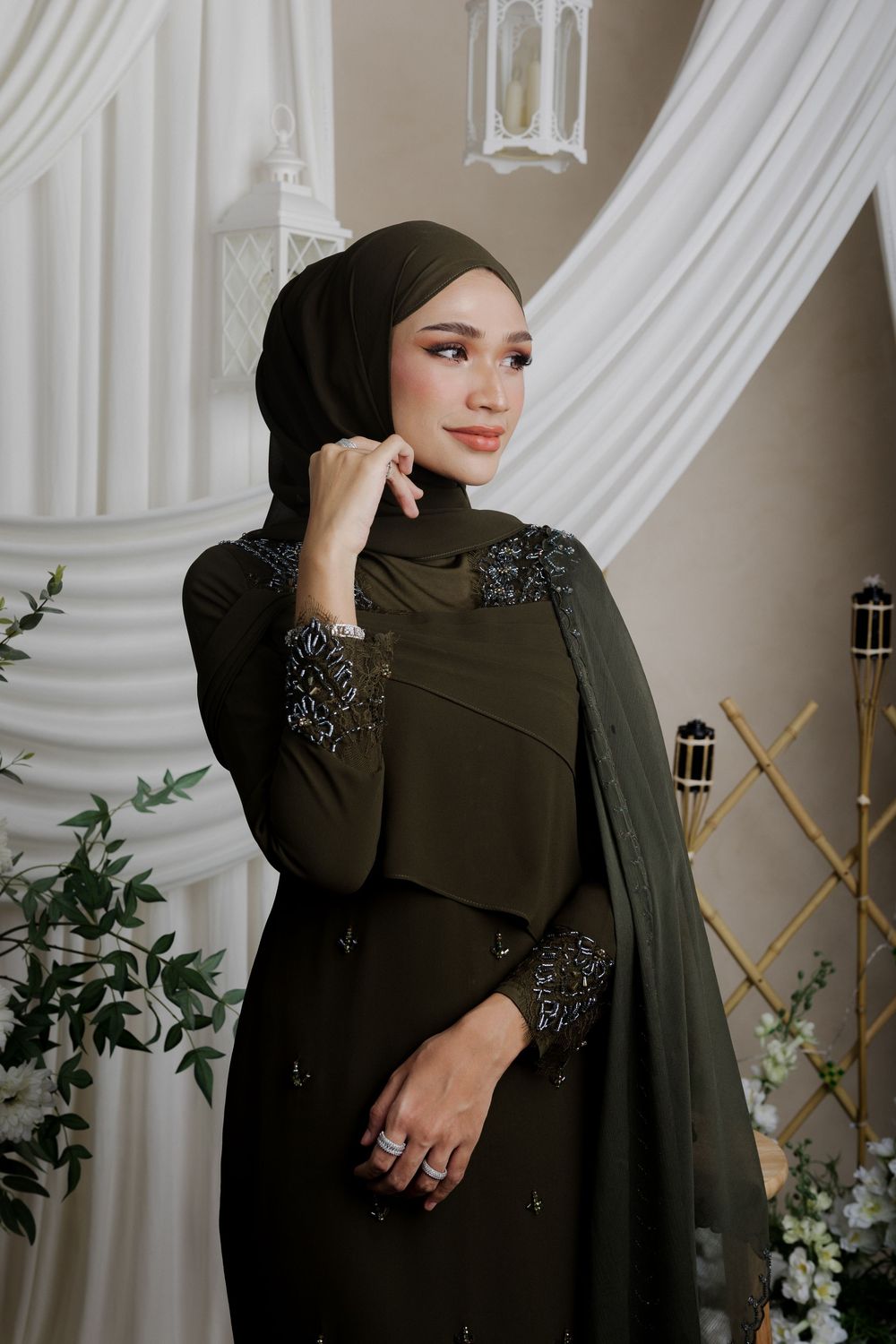 Elegant olive green abaya dress with intricate beaded details and matching hijab against white draped backdrop with lanterns.