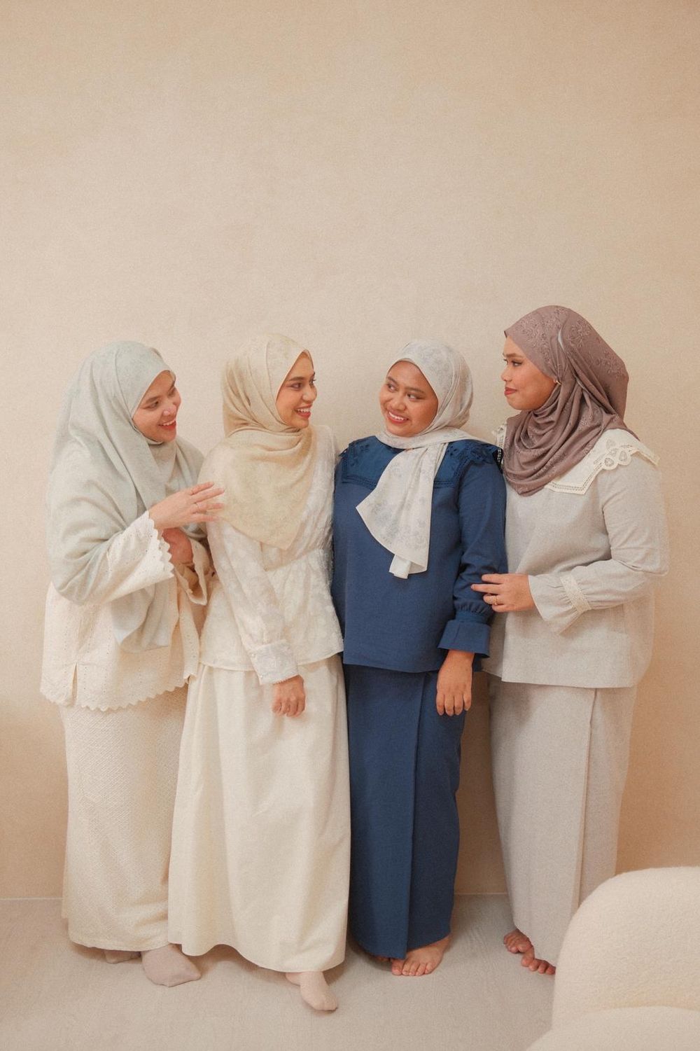 Four women wearing hijabs and modest outfits in neutral and blue tones, smiling and posing together indoors.