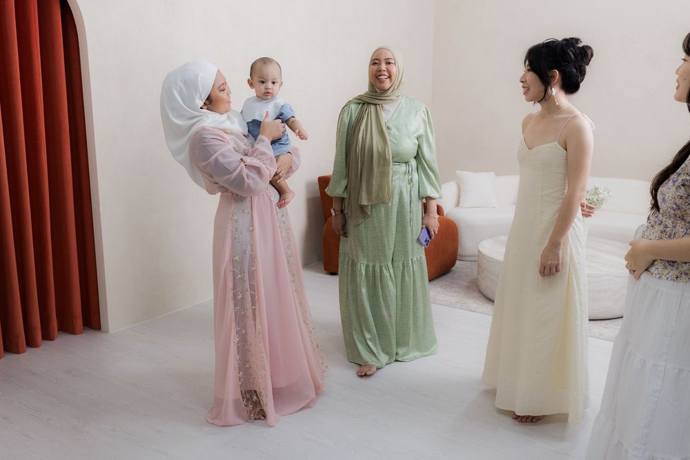 People in pastel pink and green dresses gather in an elegant room with burgundy curtains during a formal event.
