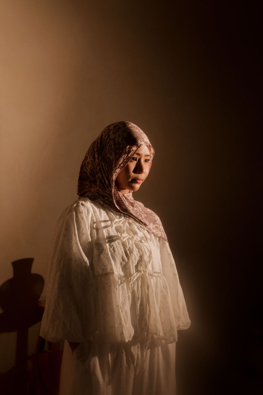 A young woman in a white lace dress and floral hijab stands in warm dramatic lighting against a dark background.