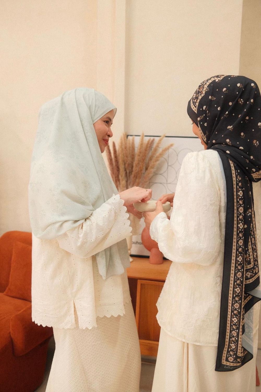 Two Muslim women in hijabs and white outfits exchanging a gift in a warmly decorated living room.