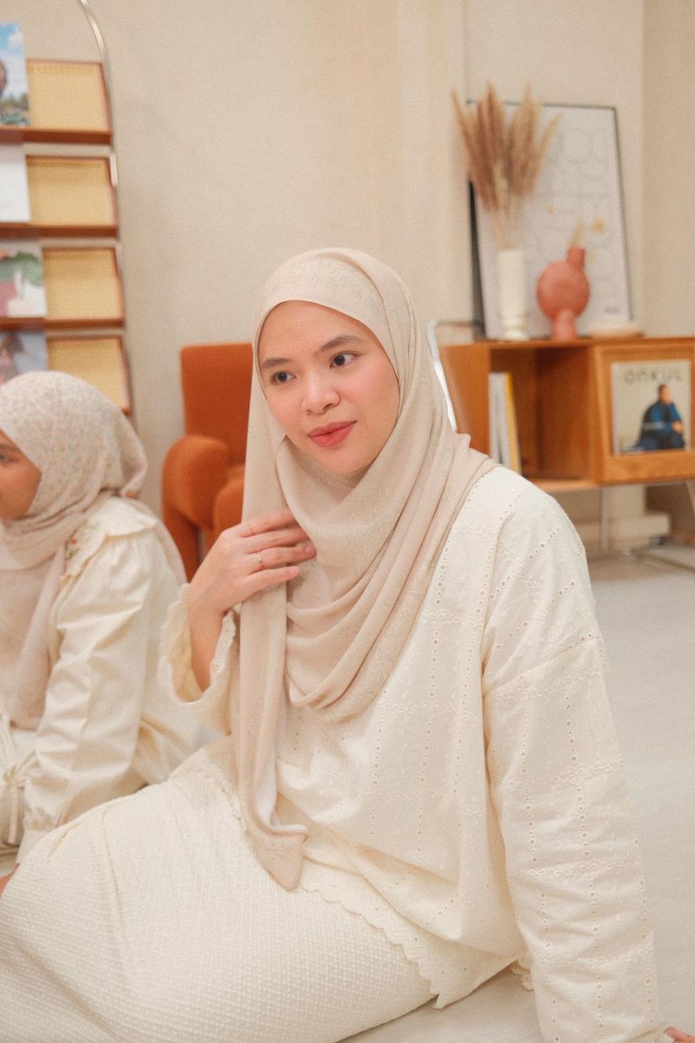 A woman wearing a cream hijab and white outfit sits gracefully in a warmly decorated room.