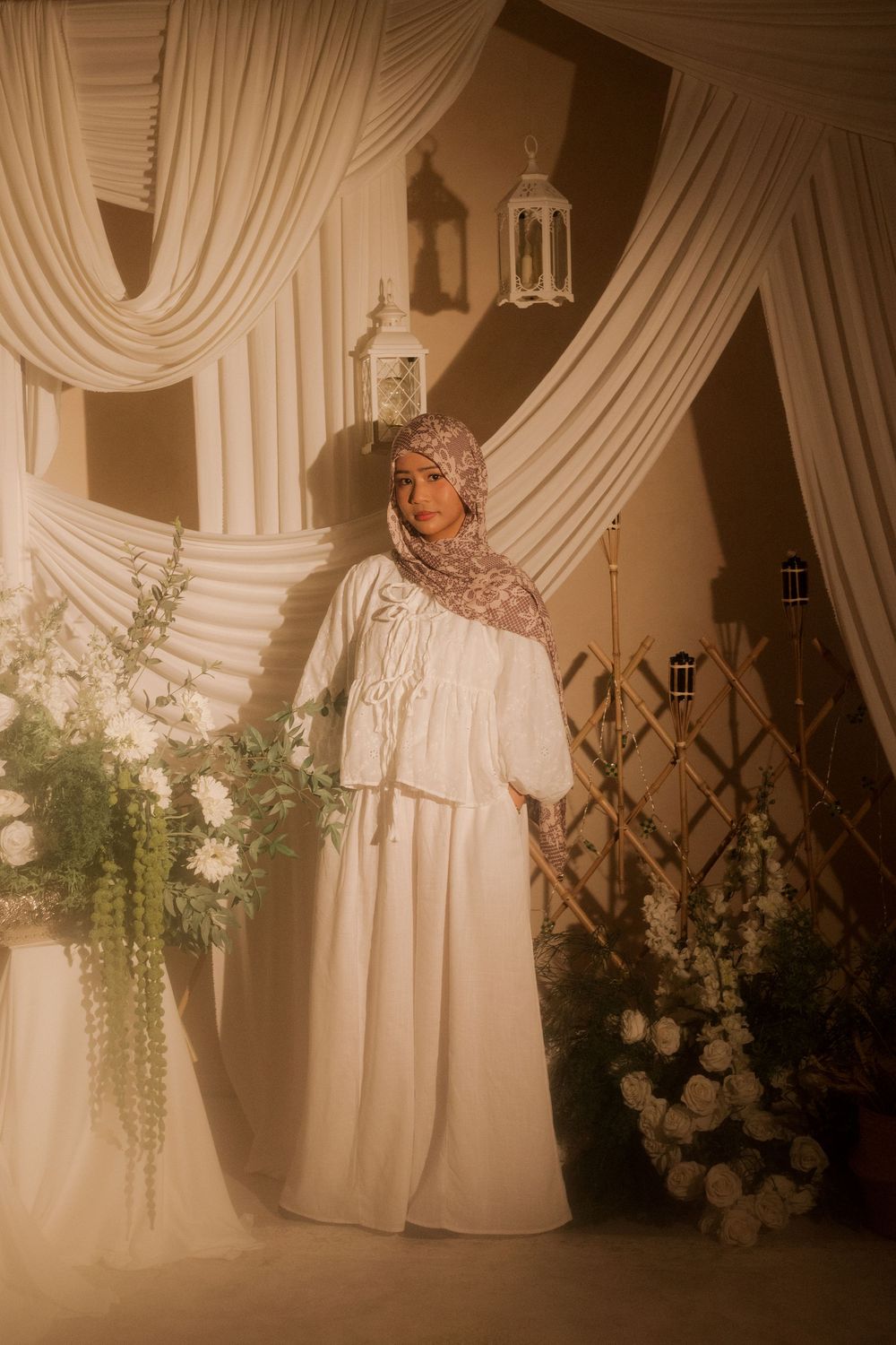 A woman in a white modest dress and patterned hijab poses gracefully among white flowers and draped fabric backdrop.