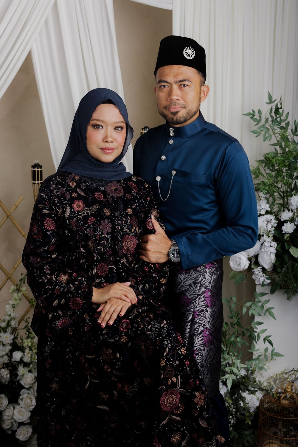 A couple in traditional Malay formal attire poses together against a white floral backdrop for a special occasion.