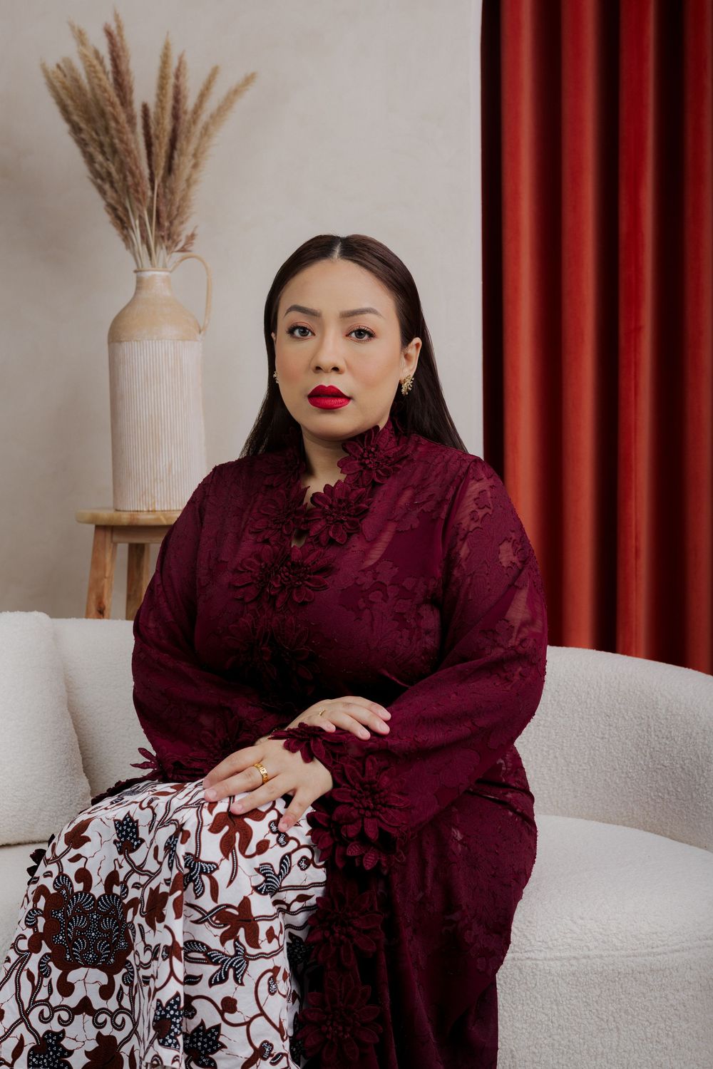 Woman in burgundy lace top and floral skirt sits elegantly on white couch with pampas grass decor and red curtain.