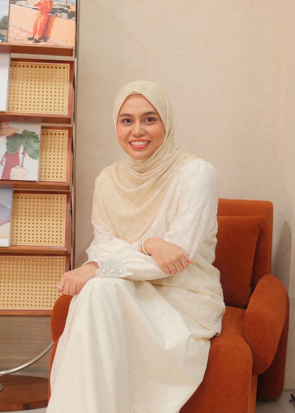Smiling woman in cream hijab and white outfit seated in an orange chair near a wooden bookshelf.