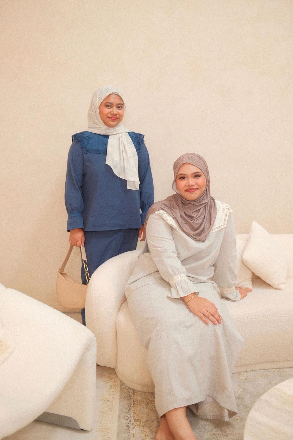 Two women wearing hijabs pose together, one in blue outfit standing, another in gray seated on white sofa.