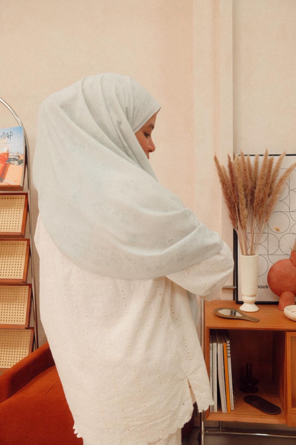 Woman wearing a white hijab and white lace dress standing in a stylish room with dried pampas grass decor.