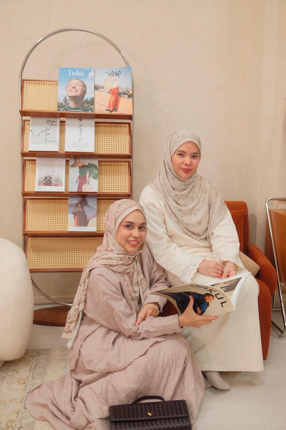 Two women in hijabs pose near a magazine rack, one seated on floor, other on chair holding a magazine.