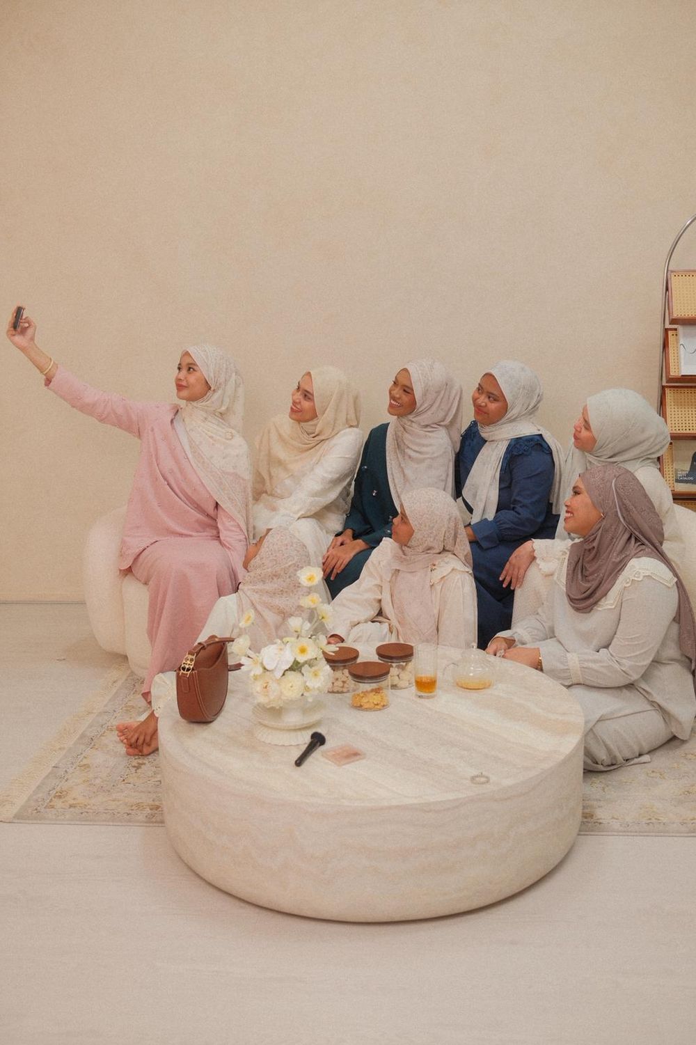 Group of Muslim women in hijabs taking a selfie together around a marble coffee table in a bright room.