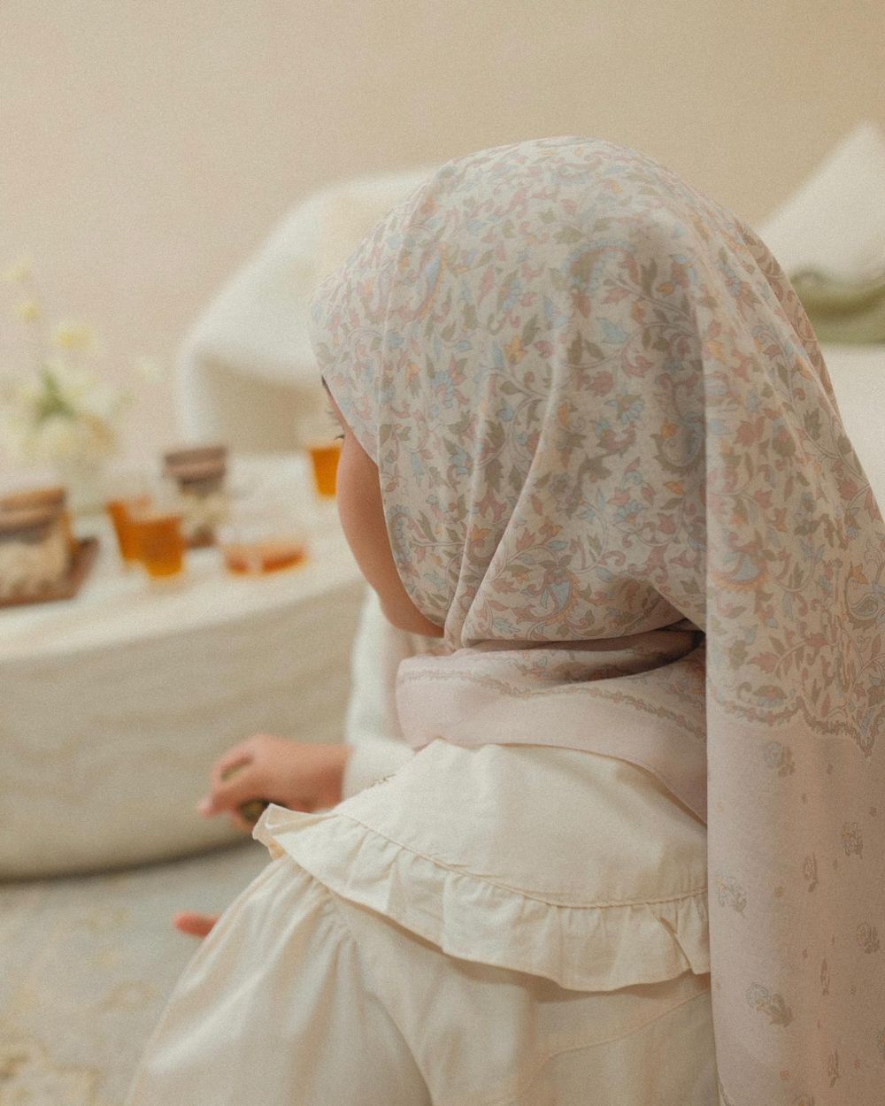 Young child in floral hijab and white ruffled dress sits at a low table with tea cups and flowers.