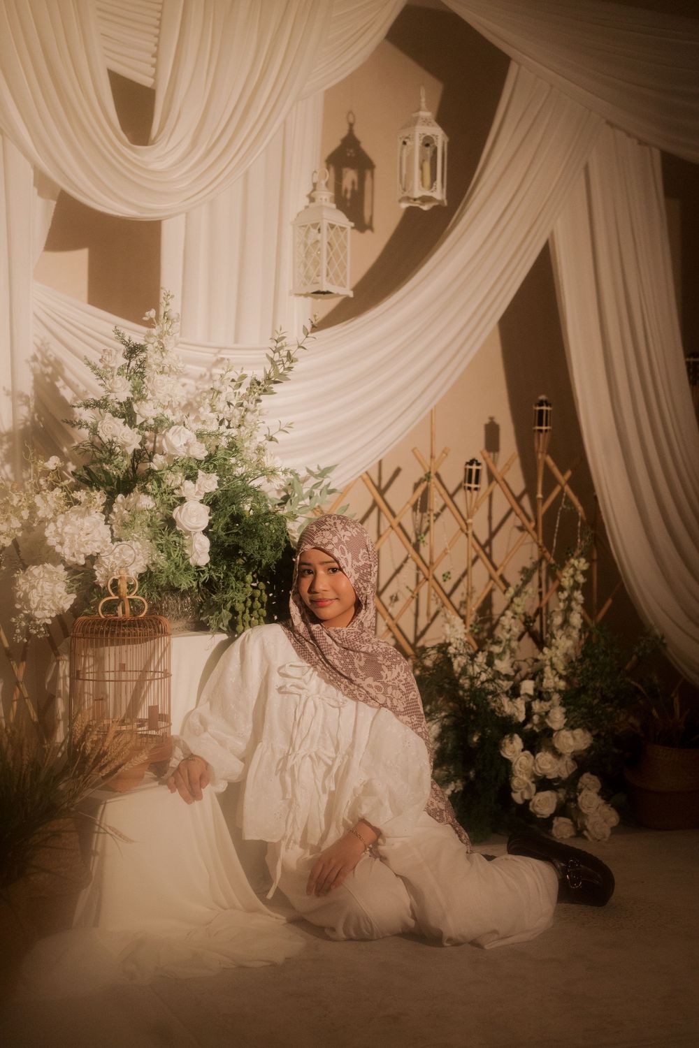 Woman in hijab and white outfit sits gracefully among white flowers and elegant draping decor.
