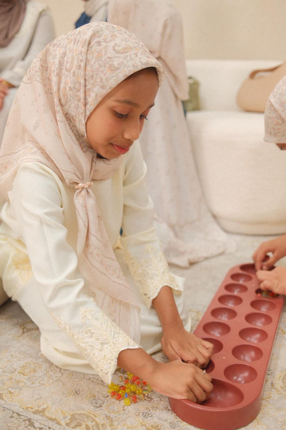 Young Muslim girl in hijab playing mancala board game on floor during Eid celebration.