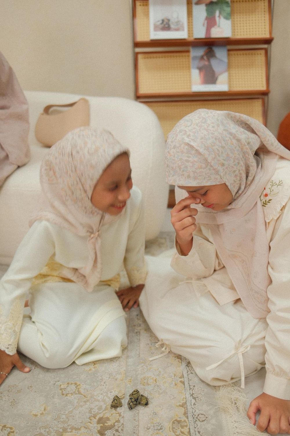 Two young girls in white outfits and floral hijabs sit together on a rug, sharing a sweet moment.