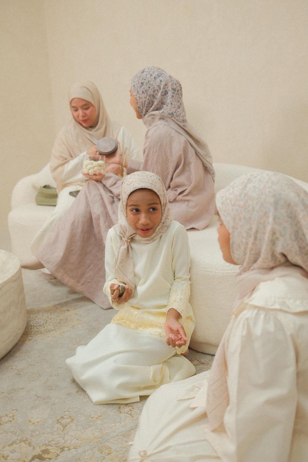 Muslim women and girls in hijabs and traditional attire sit together in a bright, cozy room during a gathering.