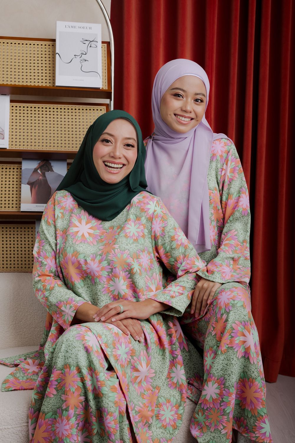 Three people wearing matching floral dresses and hijabs in pink and green colors pose together on a couch.