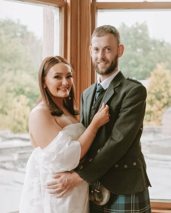 Couple shares tender moment by window in traditional Scottish wedding attire.