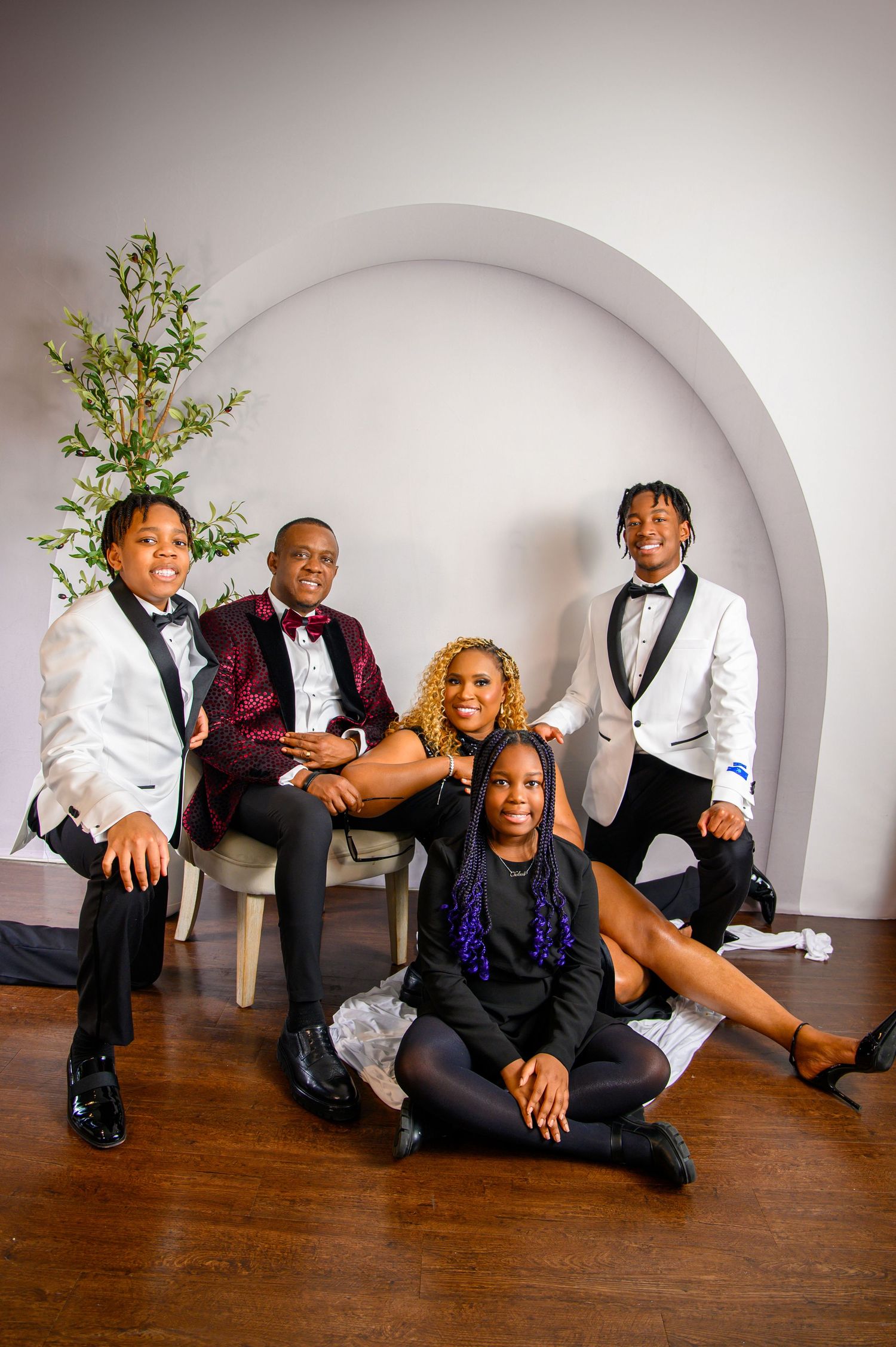 A group in formal attire poses playfully on a hardwood floor against a white curved wall.