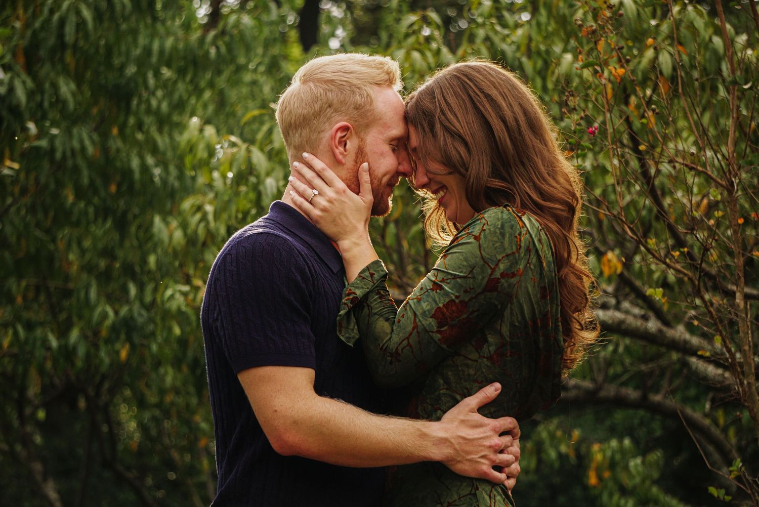 A couple shares a romantic kiss in front of lush green foliage in an outdoor setting.