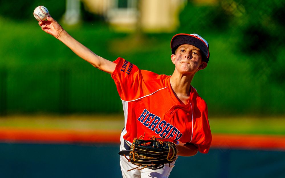 Young baseball pitcher in orange jersey throwing from the mound, SportsFox Photography.