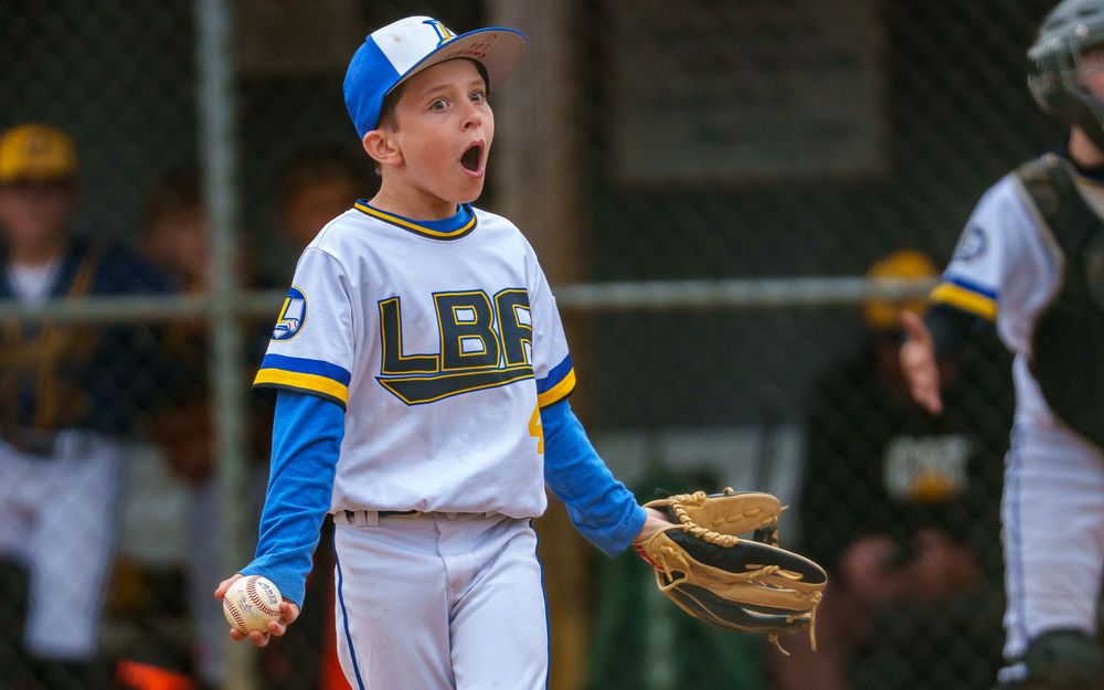 Little league player in blue and white uniform smiling in the dugout, SportsFox Photography.