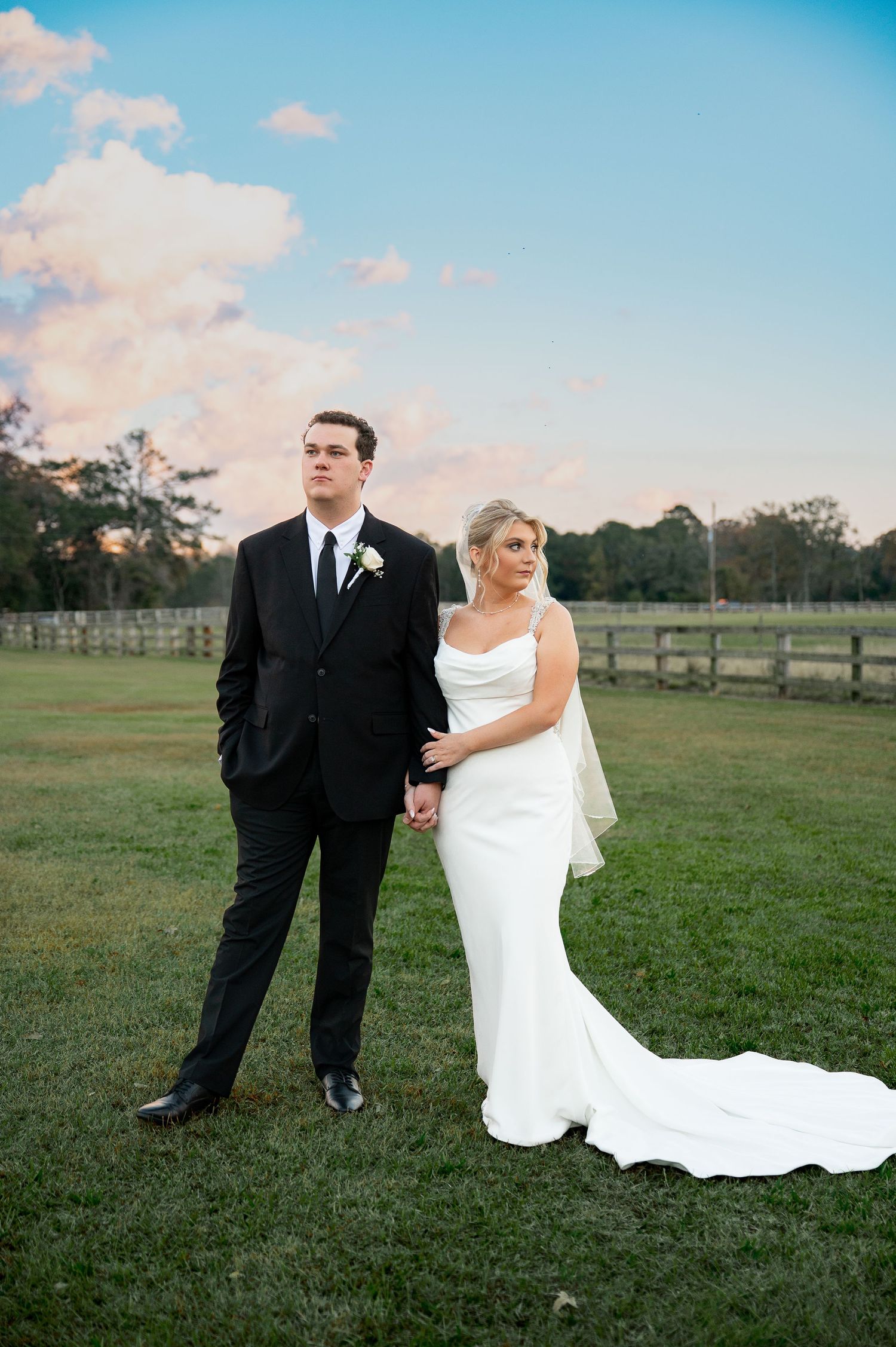 Elegant couple poses in formal wedding attire against sunset sky and rustic fence.