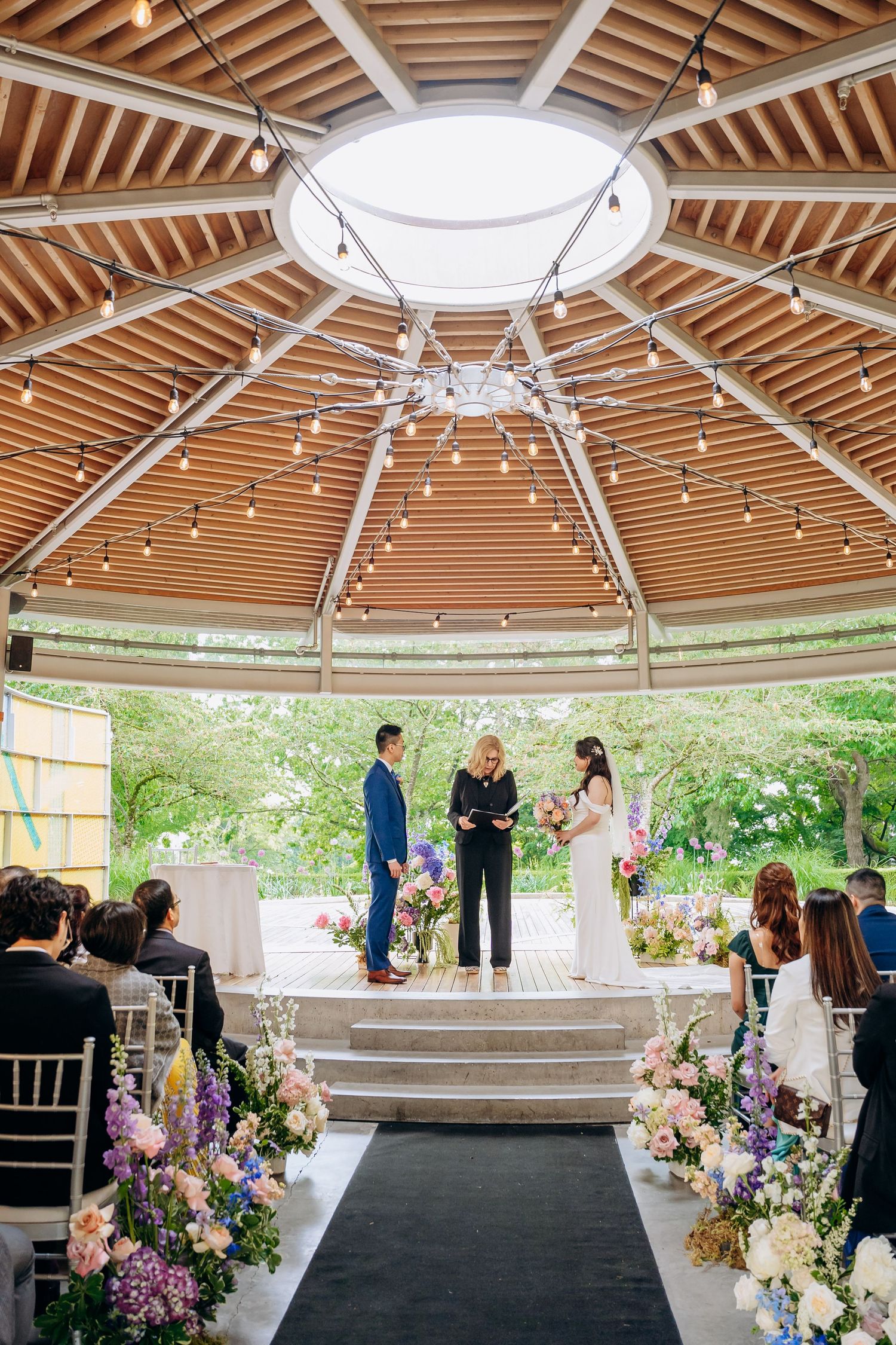 Panoramic view of an outdoor wedding ceremony setup under a wooden gazebo with floral arrangements and black aisle runner.