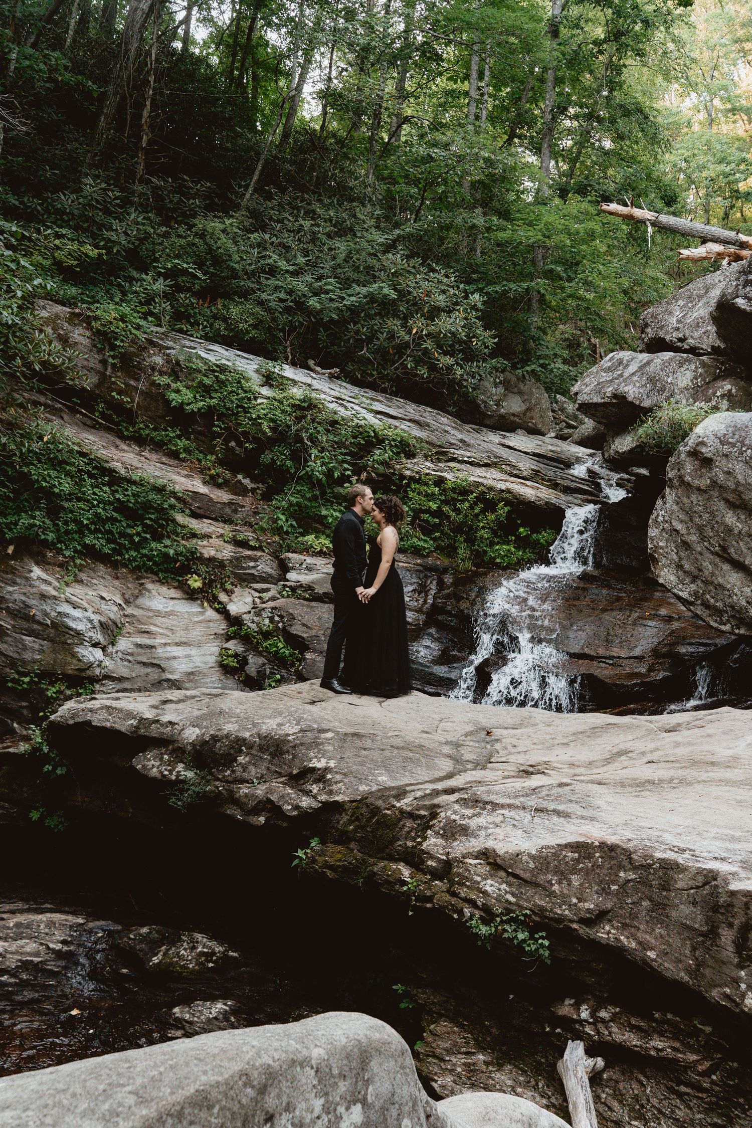 A couple embraces while sitting in a natural pool beneath a rocky cliff with a cascading waterfall in the background.