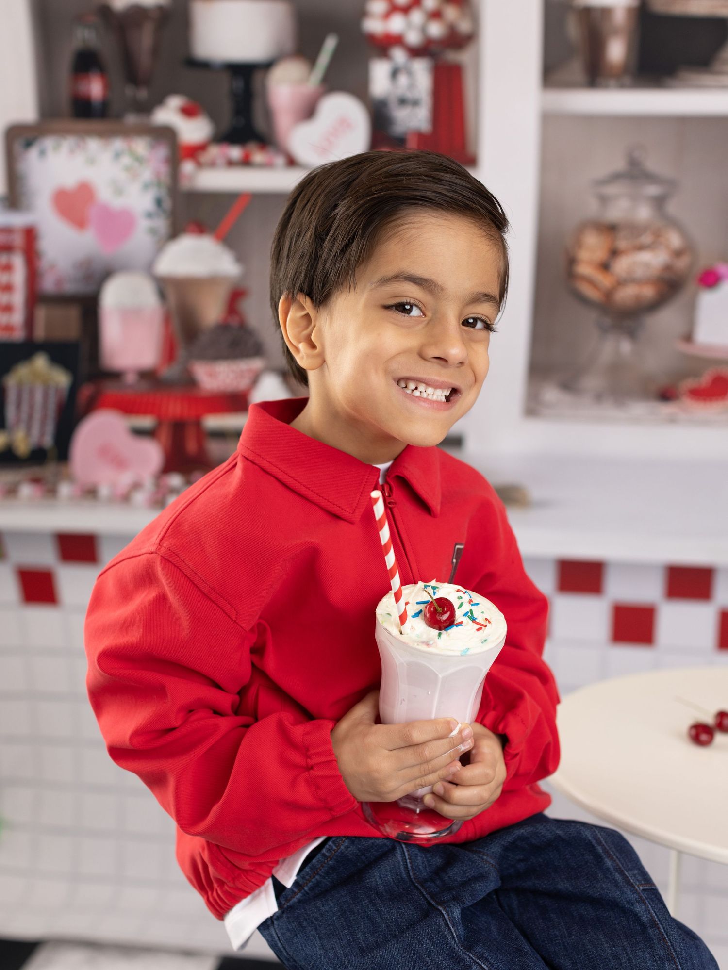 Child in red sweater enjoys a festive holiday milkshake topped with whipped cream and cherry in retro diner setting.