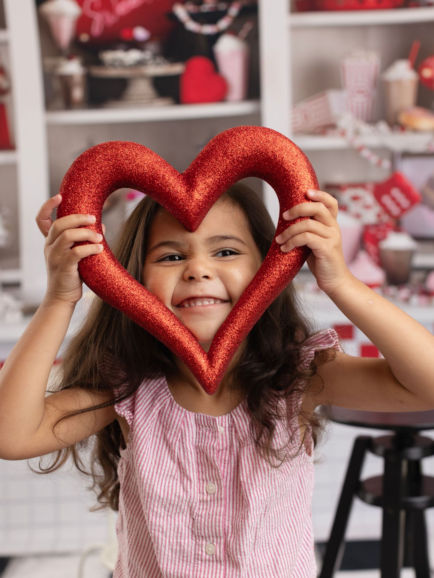 A smiling child holds up a glittery red heart decoration against a blurred background with Valentine's decor.