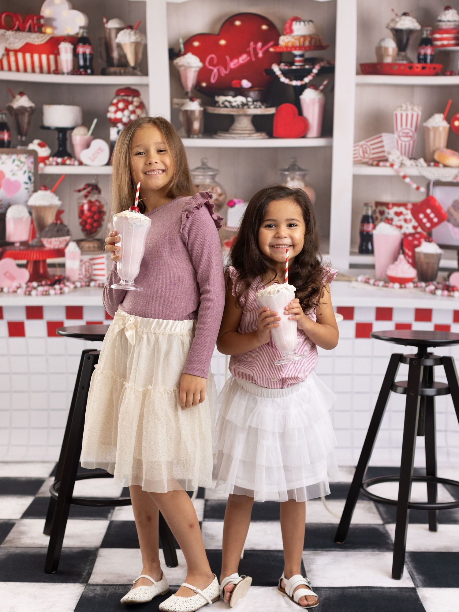 Two children in tulle skirts hold milkshakes in front of a retro diner-style display with red and white decor.