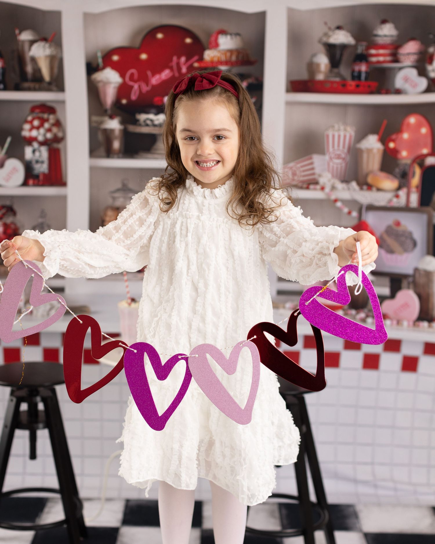 A child in a white lace dress holds a pink paper garland in a red and white decorated room.
